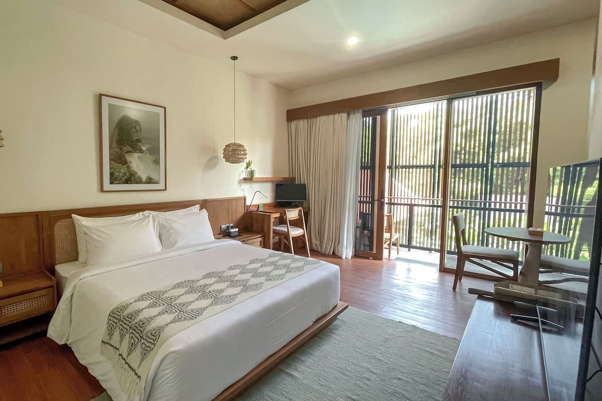 A spacious bedroom with a large bed, a desk, and a chair, a television, and a balcony with a view of greenery.