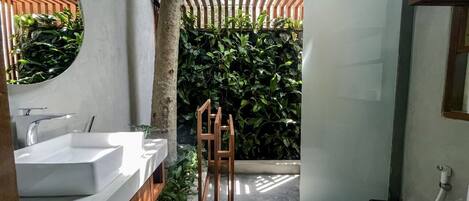 Une salle de bain avec un lavabo blanc, un miroir et une douche. Un tabouret en bois et une plante en pot se trouvent dans un coin.