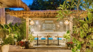 An outdoor seating area with three tables and chairs, surrounded by potted plants and a thatched roof structure. - Leafy Home Canggu (Canggu)