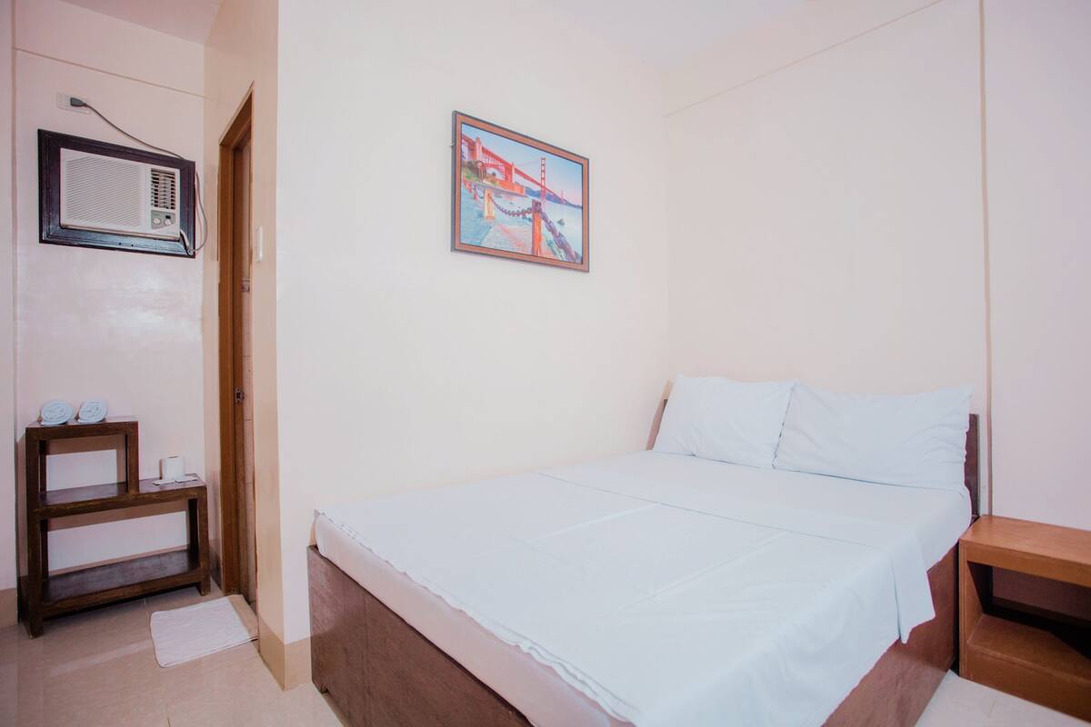 Un dormitorio con cama, una mesita de noche, un estante pequeño, un aire acondicionado de pared y un cuadro en la pared.