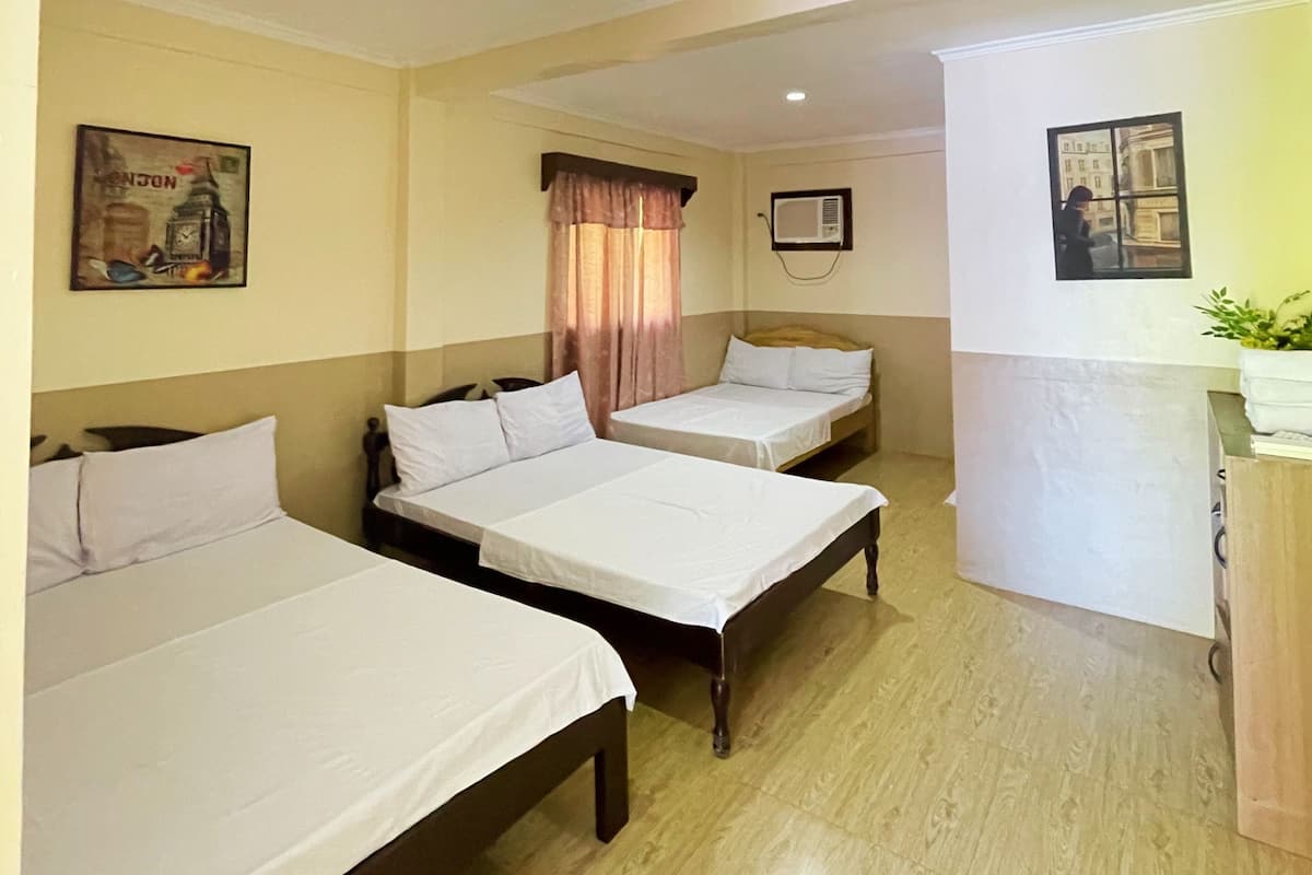 Habitación de hotel con tres camas individuales, cabecera de madera, ventana con cortinas, equipo de aire acondicionado y un cuadro en la pared.