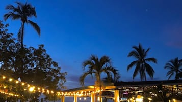 An outdoor dining area with string lights, palm trees, and a view of the ocean at dusk.