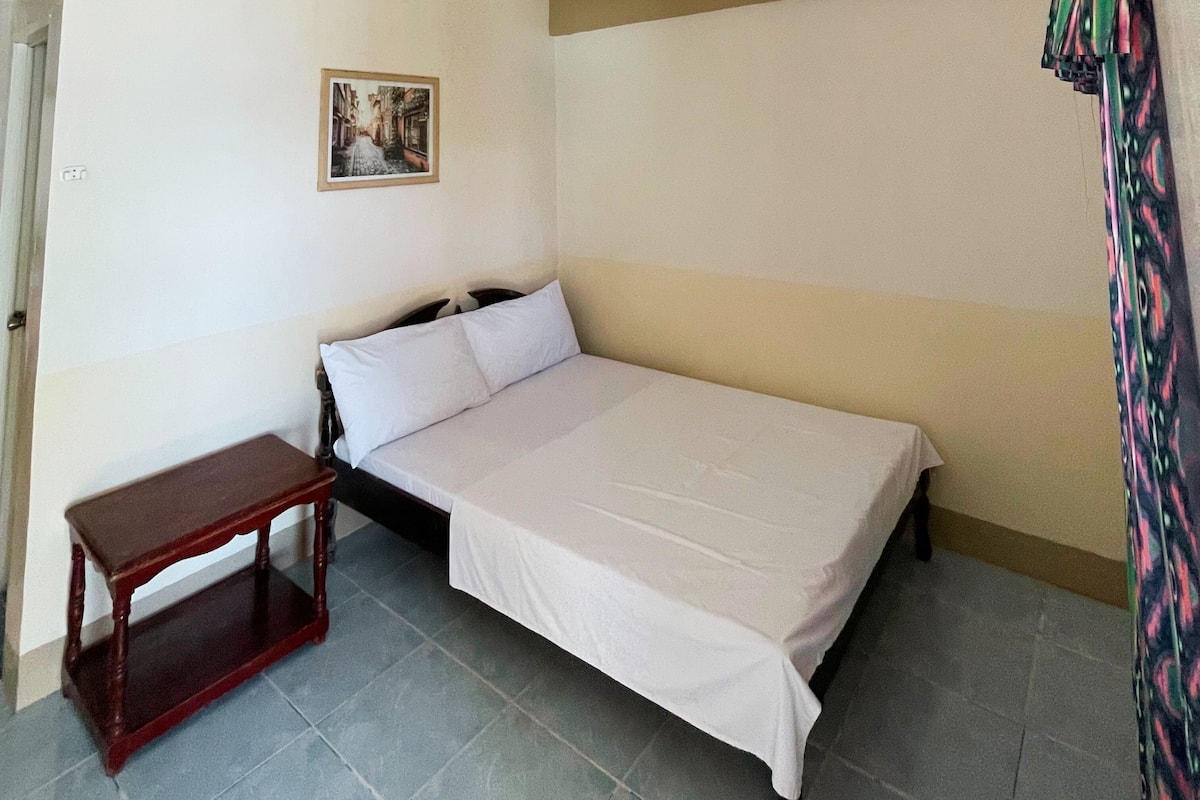 Una cama individual con sábana blanca en una habitación con una pequeña mesa de madera y un cuadro en la pared.