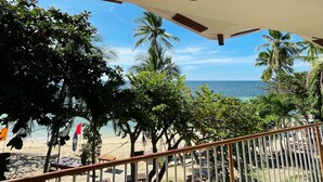 Vista a la playa desde un balcón, con palmeras y una playa de arena al fondo.