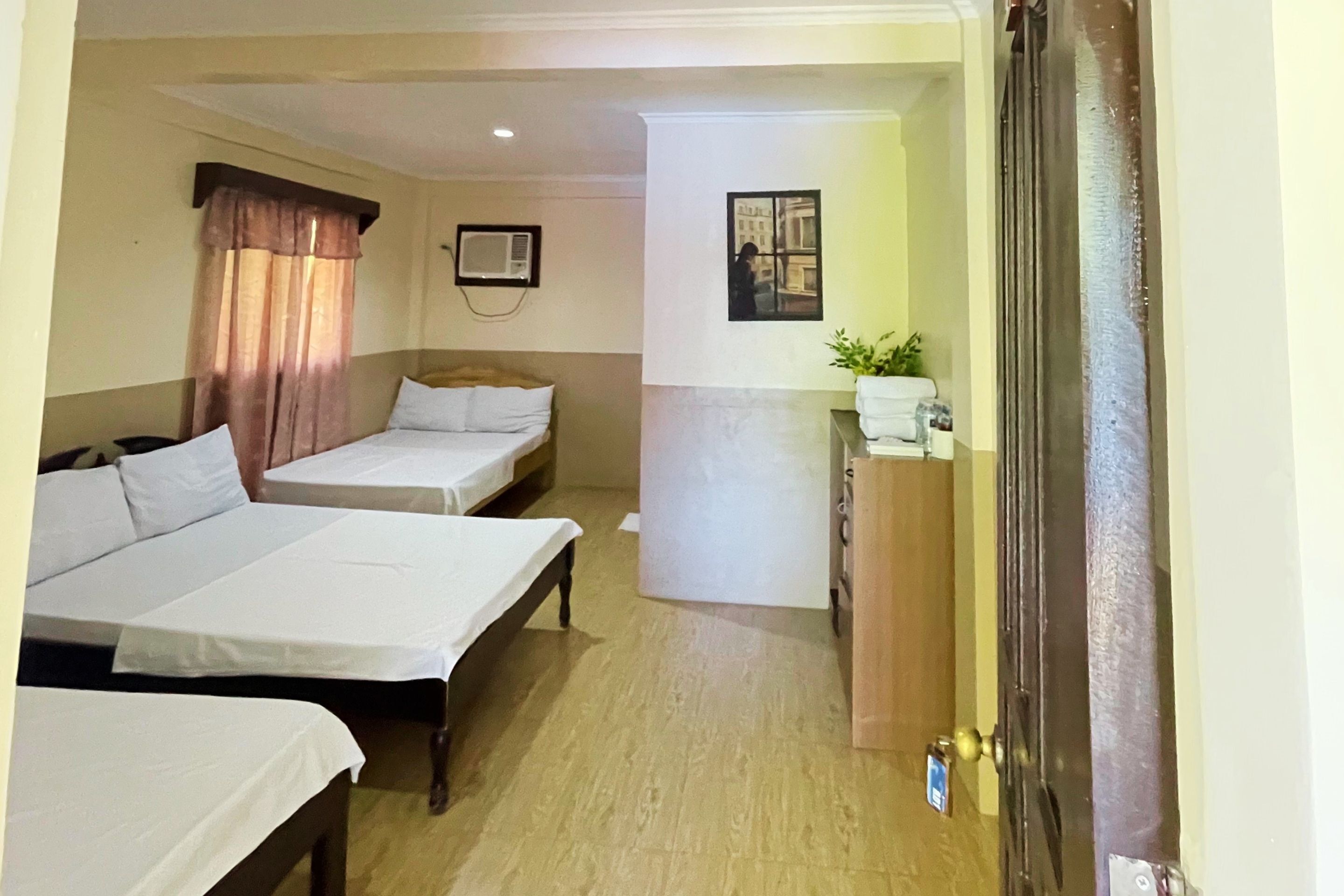 A hotel room with three single beds, a wooden floor, a small desk with a plant, and a door with a brass handle.