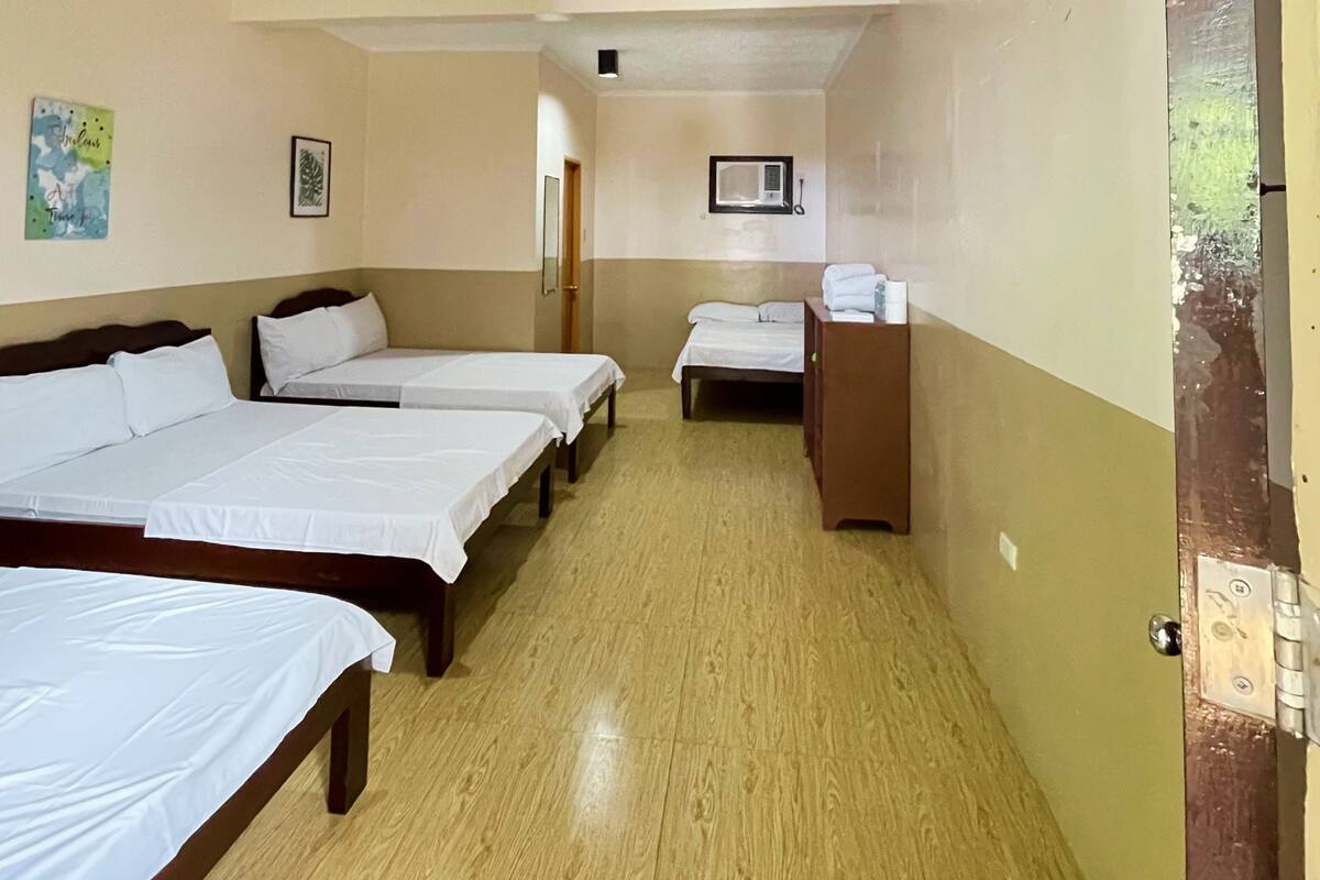Habitación de hotel con tres camas individuales, piso de madera, un escritorio pequeño con espejo y un equipo de aire acondicionado instalado en la pared.