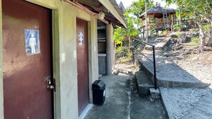 Un edificio con una puerta marrón señalada para uso exclusivo de hombres, un pequeño espacio al aire libre con escalones que conducen a una estructura con techo de paja, y un bote de basura negro cerca de la entrada.