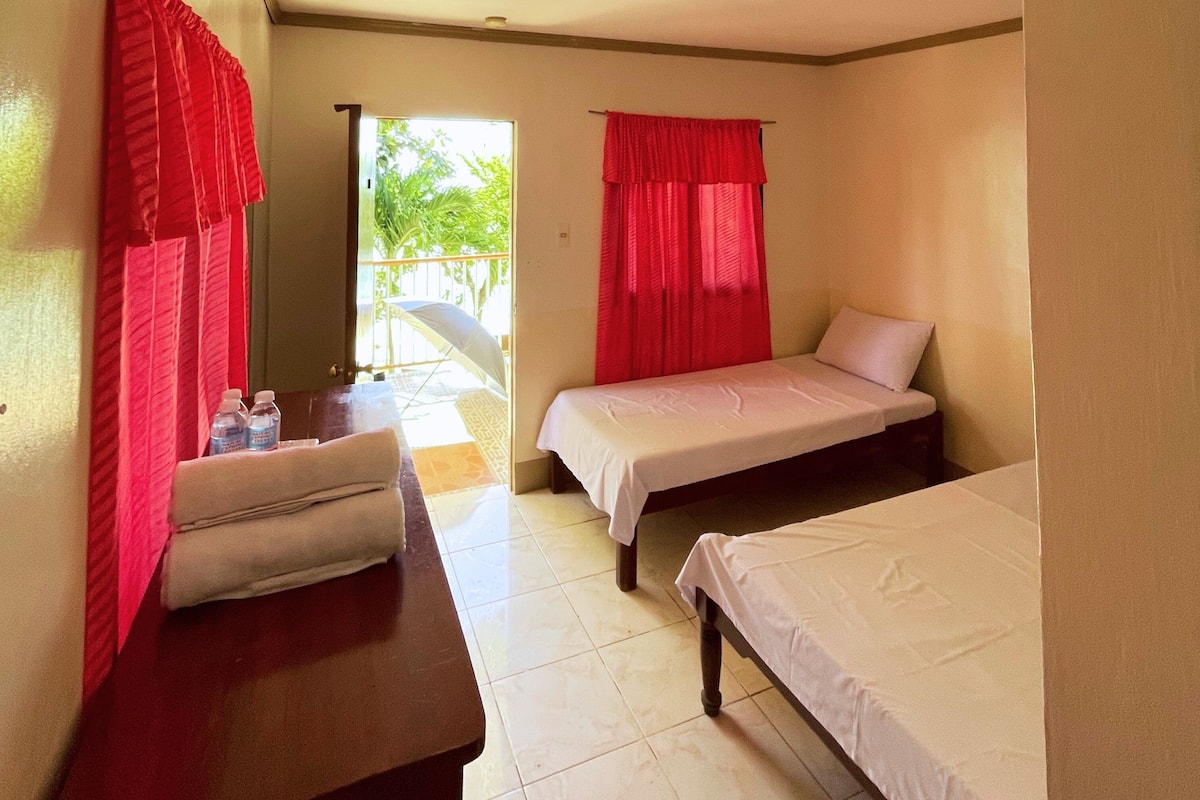 Habitación de hotel con dos camas individuales, una mesita con una silla y un balcón con cortinas rojas.
