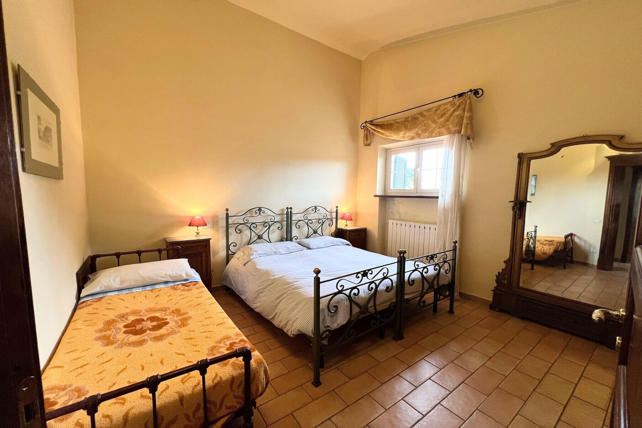 A bedroom with a single bed, a bedside table with a lamp, a window with a curtain, and a mirror.