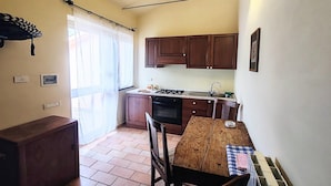 A compact kitchen with wooden cabinets, a stove, and a sink. There is a dining table with chairs and a window with sheer curtains.
