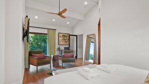 A spacious bedroom with a large bed, a ceiling fan, and a view of greenery through the window.