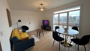 Ein modernes Wohnzimmer mit einer Couch, einem Esstisch mit Stühlen, einem Fernseher und einem großen Fenster mit Blick auf die Stadt.