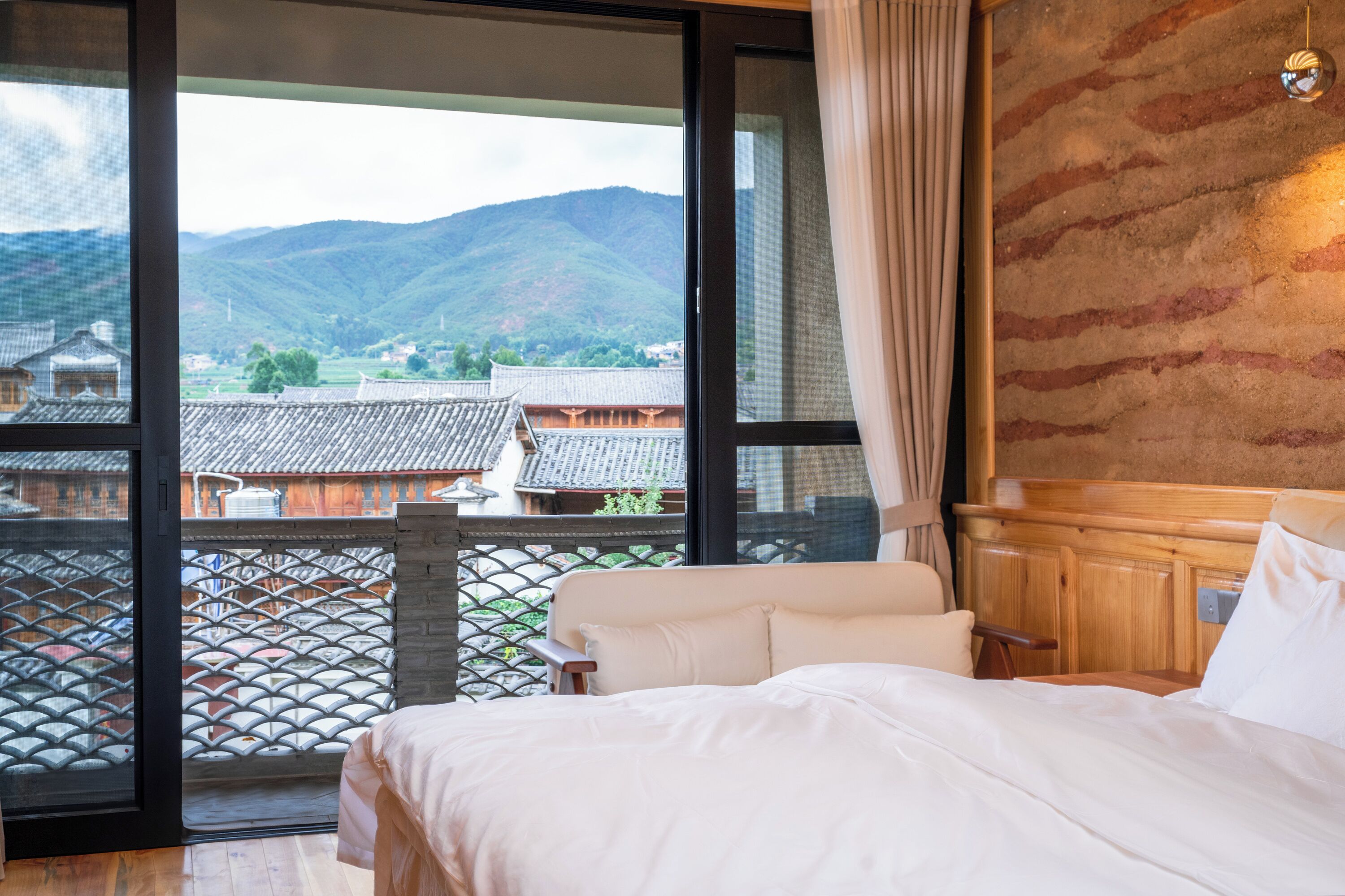 A bedroom with a large window, a bed, and a view of traditional buildings and mountains.