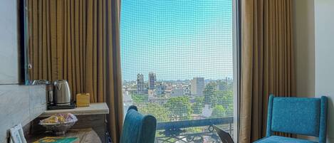A room with a large window, a laptop on a small table, a blue chair, and a bowl of fruit on a shelf.