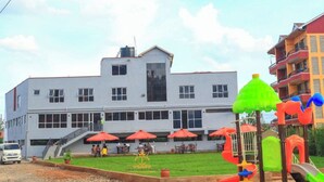 A modern hotel with a flat roof, multiple windows, and a central staircase. There are outdoor seating areas with red umbrellas and a playground with colorful slides and play structures. - Tawala Garden Hotel (Nairobi)