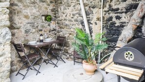 Une terrasse extérieure avec un mur en pierre, une table avec des chaises, un pot de fleurs et un barbecue.