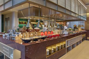 Buffet montado com diversos alimentos e utensílios, um chefe preparando comida e interior bem iluminado.