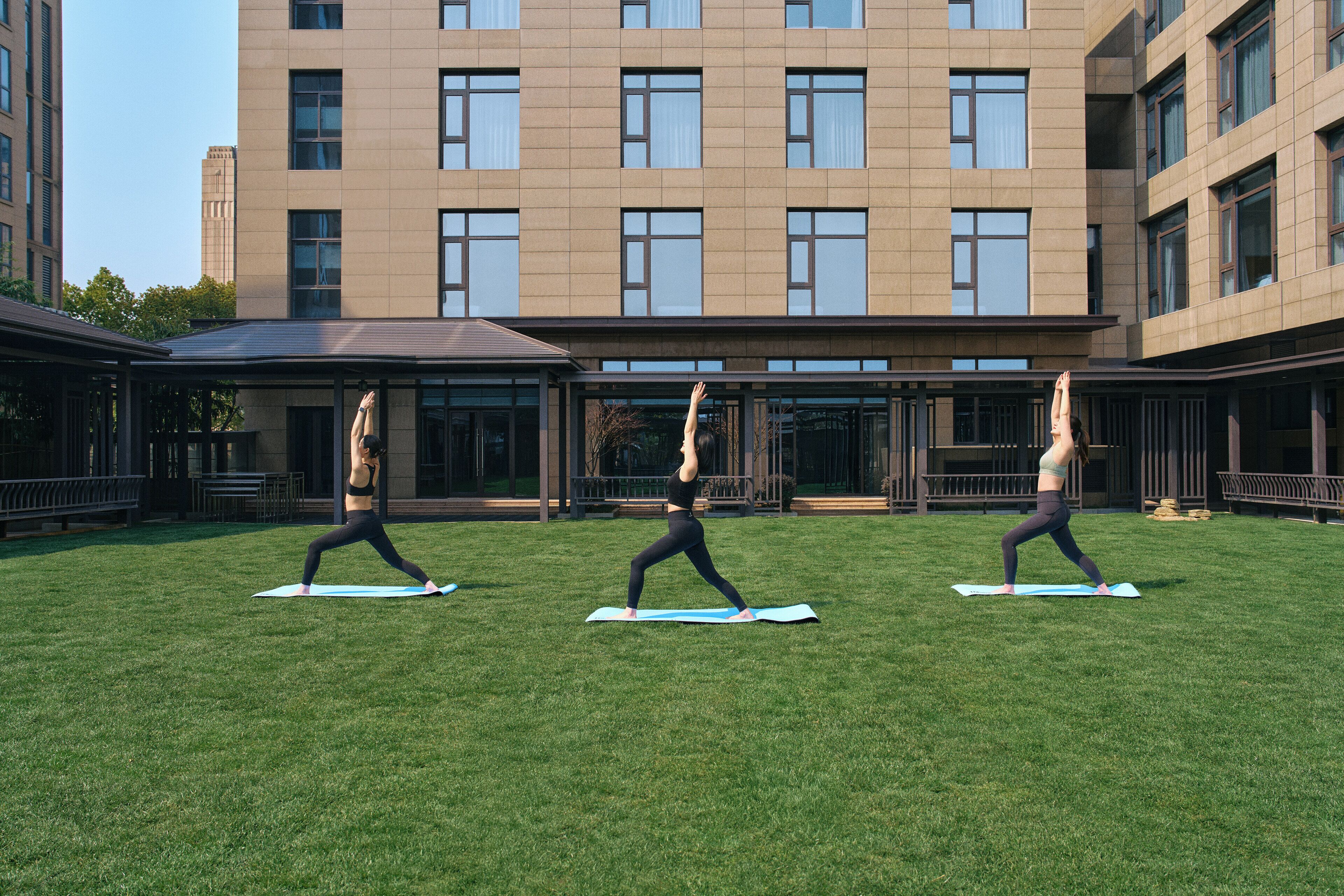 A courtyard with three people doing yoga on mats, surrounded by a building with large windows and a flat roof.