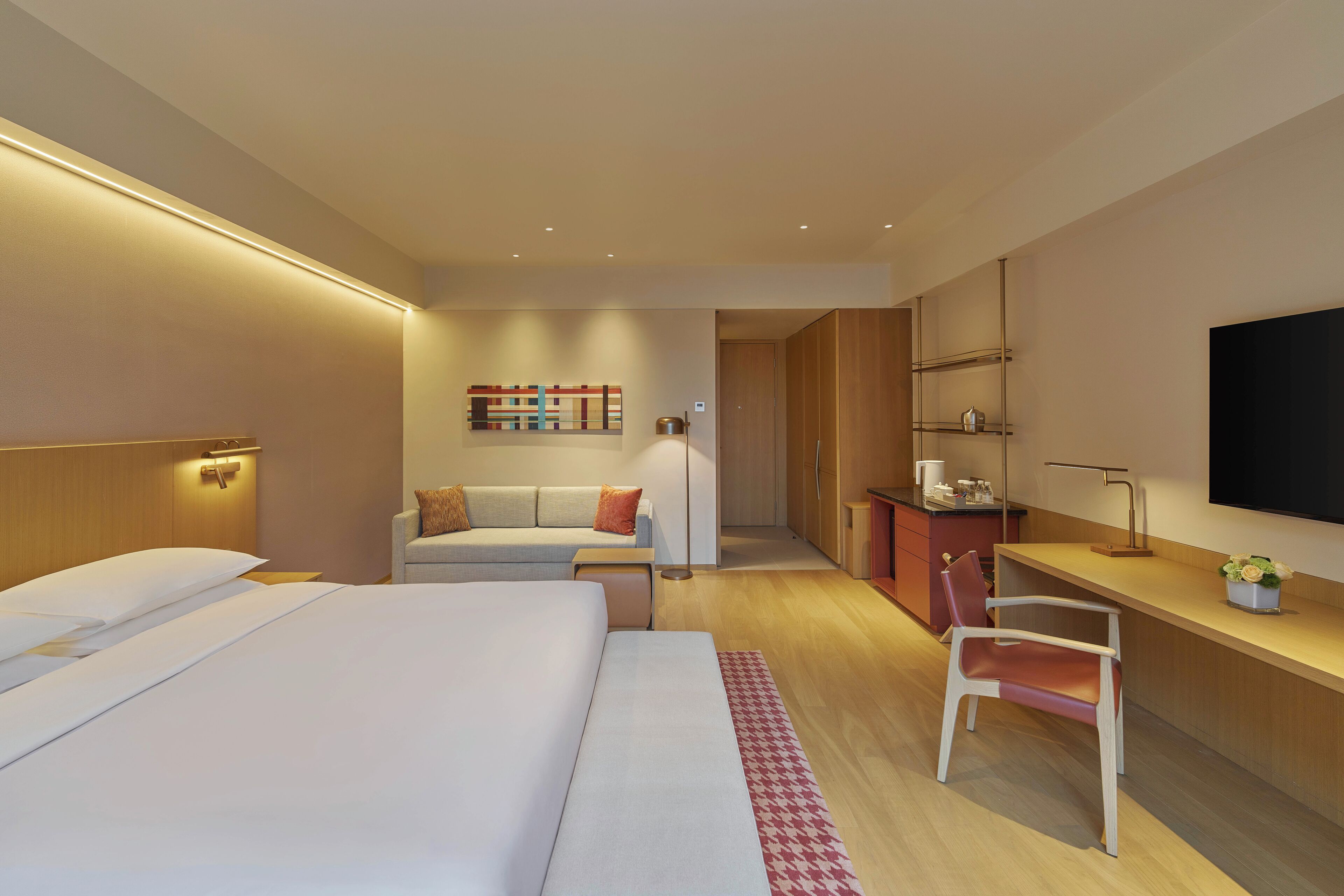 A modern hotel room with a large bed, a sofa, a desk, and a TV.