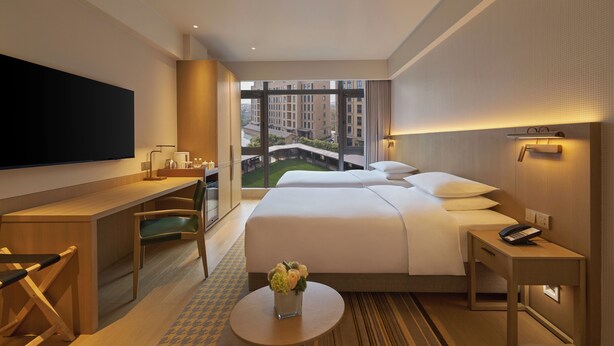 Une chambre d’hôtel avec un grand lit, un bureau, une chaise, une petite table ornée d’un vase de fleurs, un téléviseur fixé au mur, et une vue sur des immeubles et un pont, visible par la fenêtre.