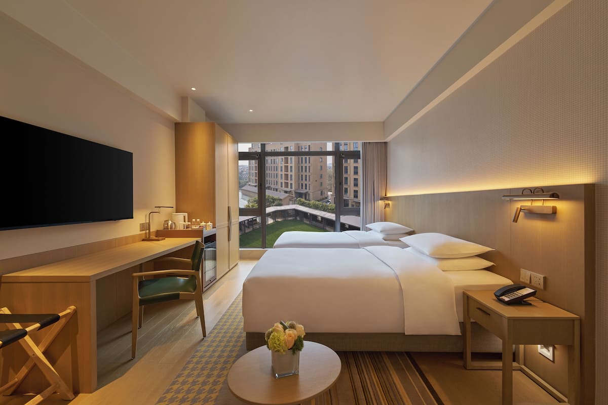 Hotelkamer met een groot bed, een bureau, een stoel, een kleine tafel met een bloevas, een aan de muur bevestigde televisie en uitzicht op gebouwen en een brug door het raam.