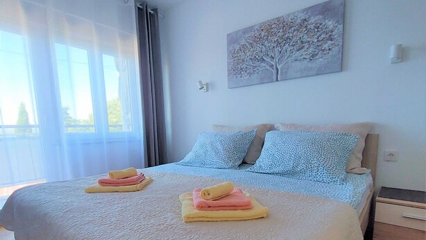 Una cama bien hecha con toallas dobladas, un cuadro de un árbol en la pared y una ventana con cortinas.