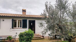 Una casa con techo de tejas, chimenea y jardín con banco, mesa y un árbol.
