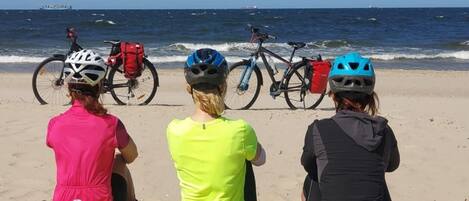 Tres personas sentadas en una playa arenosa con bicicletas y cascos, mirando hacia el océano.