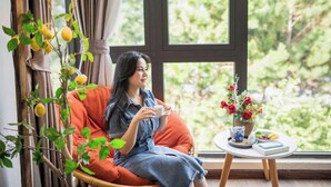 Una habitación con un ventanal grande, una silla de mimbre, una mesita con un florero y una planta con frutos amarillos.