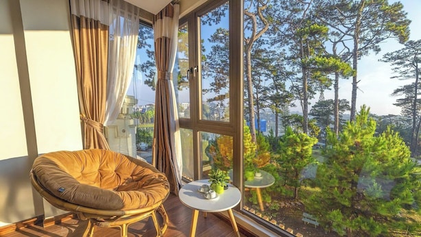 Una habitación con un ventanal grande, una silla de mimbre, una mesita redonda con una planta y vistas a árboles desde el exterior.