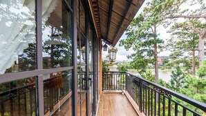 Un balcón de madera con barandillas de vidrio, vista a árboles y un edificio distante.