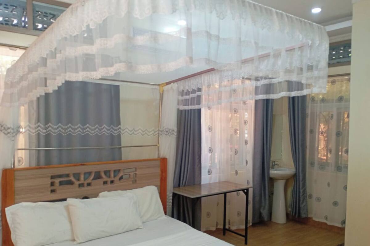 A bedroom with a bed, a bedside table, a window with curtains, and a bathroom with a sink.