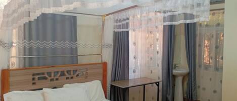A bedroom with a bed, a bedside table, a window with curtains, and a bathroom with a sink.