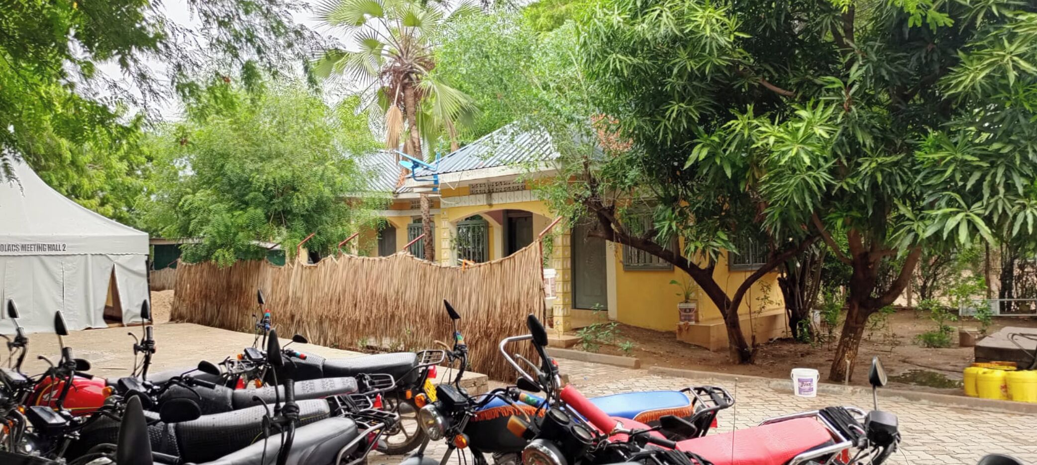 Un patio con varias motocicletas estacionadas, una carpa blanca con la leyenda "COLDS MEETING HALL 2" y un edificio de fachada amarilla.