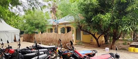 Un patio con varias motocicletas estacionadas, una carpa blanca con la leyenda "COLDS MEETING HALL 2" y un edificio de fachada amarilla.