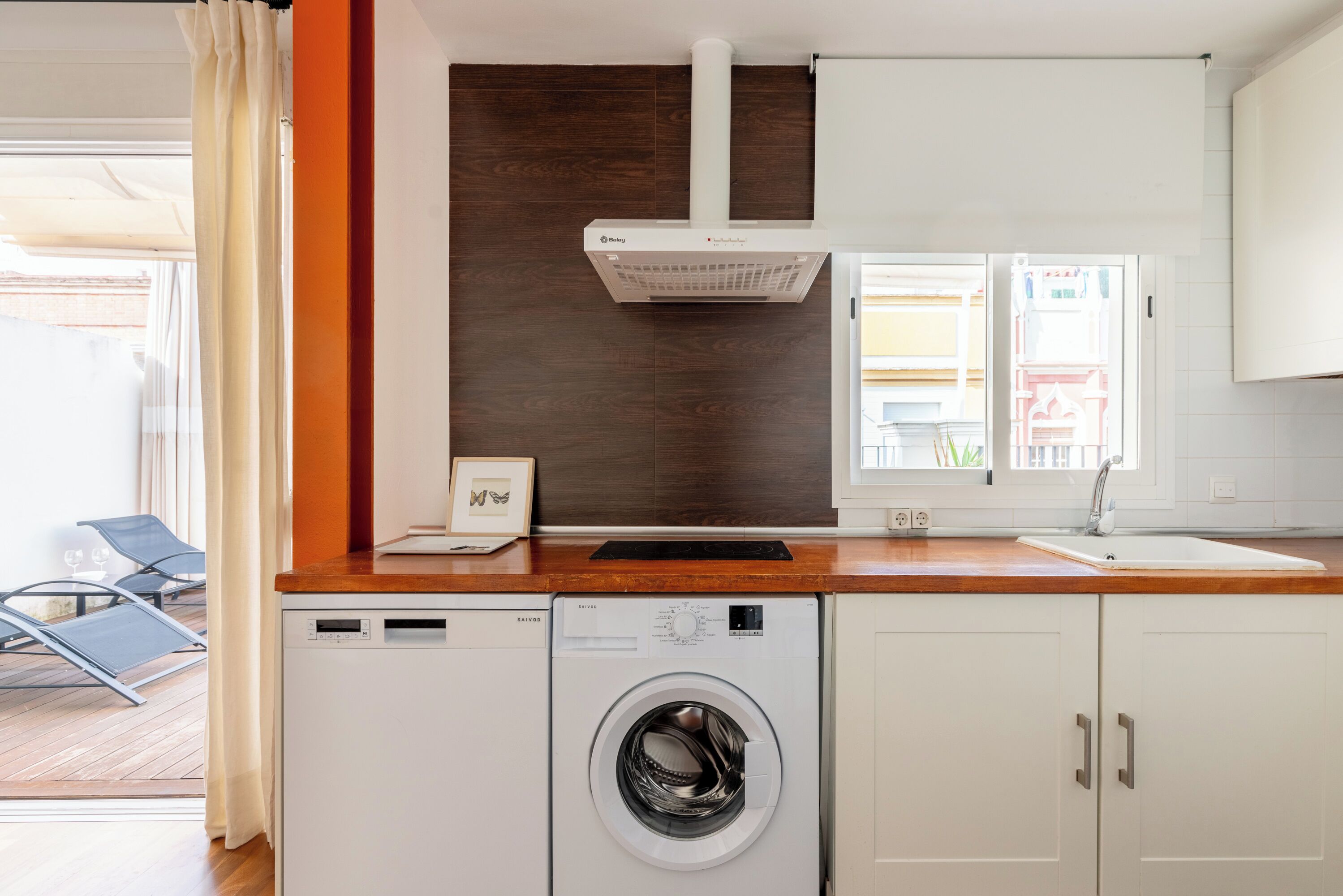 Une cuisine compacte dotée d’un lave-linge intégré, d’un plan de travail en bois et d’une fenêtre avec des rideaux.