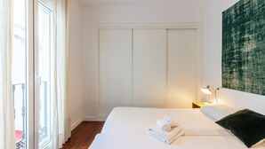 A hotel room with a bed, a nightstand with a lamp, a large window with curtains, and a green abstract painting on the wall.