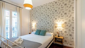 A neatly made bed with a white and gray striped blanket, two teal pillows, and a folded white towel. There are two bedside lamps on wooden nightstands, one with a vase of dried flowers. The room has a patterned wallpaper with tropical and bird motifs.