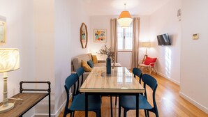A modern living room with a glass-top dining table, blue chairs, a sofa with cushions, a vase with flowers, a hanging orange pendant light, and a flat-screen TV mounted on the wall.