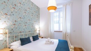 A neatly made bed with white linens and a blue blanket, a white metal headboard, and a wall with a map-themed wallpaper.