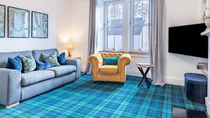 A living room with a blue plaid carpet, a grey sofa, a yellow armchair, a small round table, and a TV on the wall. - Thistle Do Nessly, Inverness (Inverness)