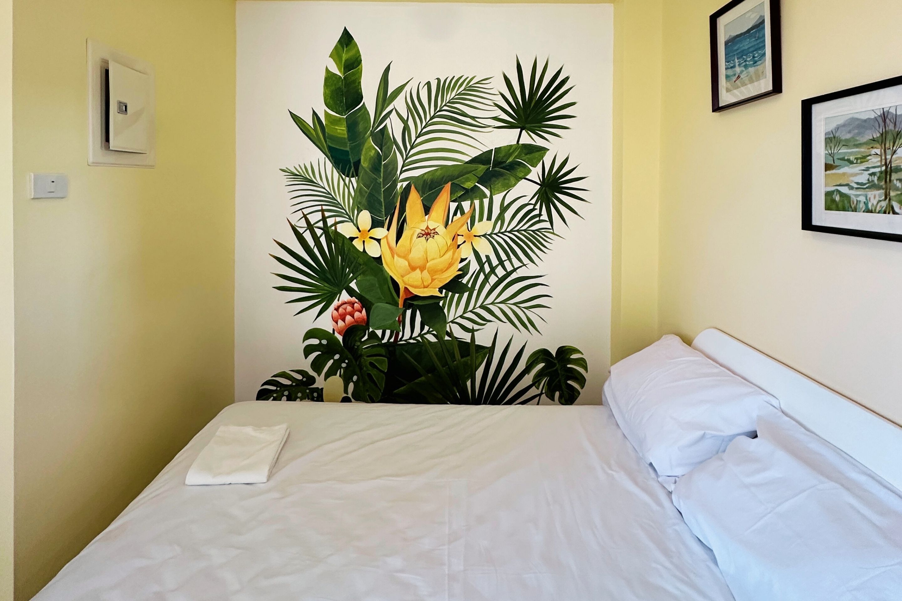 A bed with white linens, a wall-mounted light switch, and a framed picture above the bed.