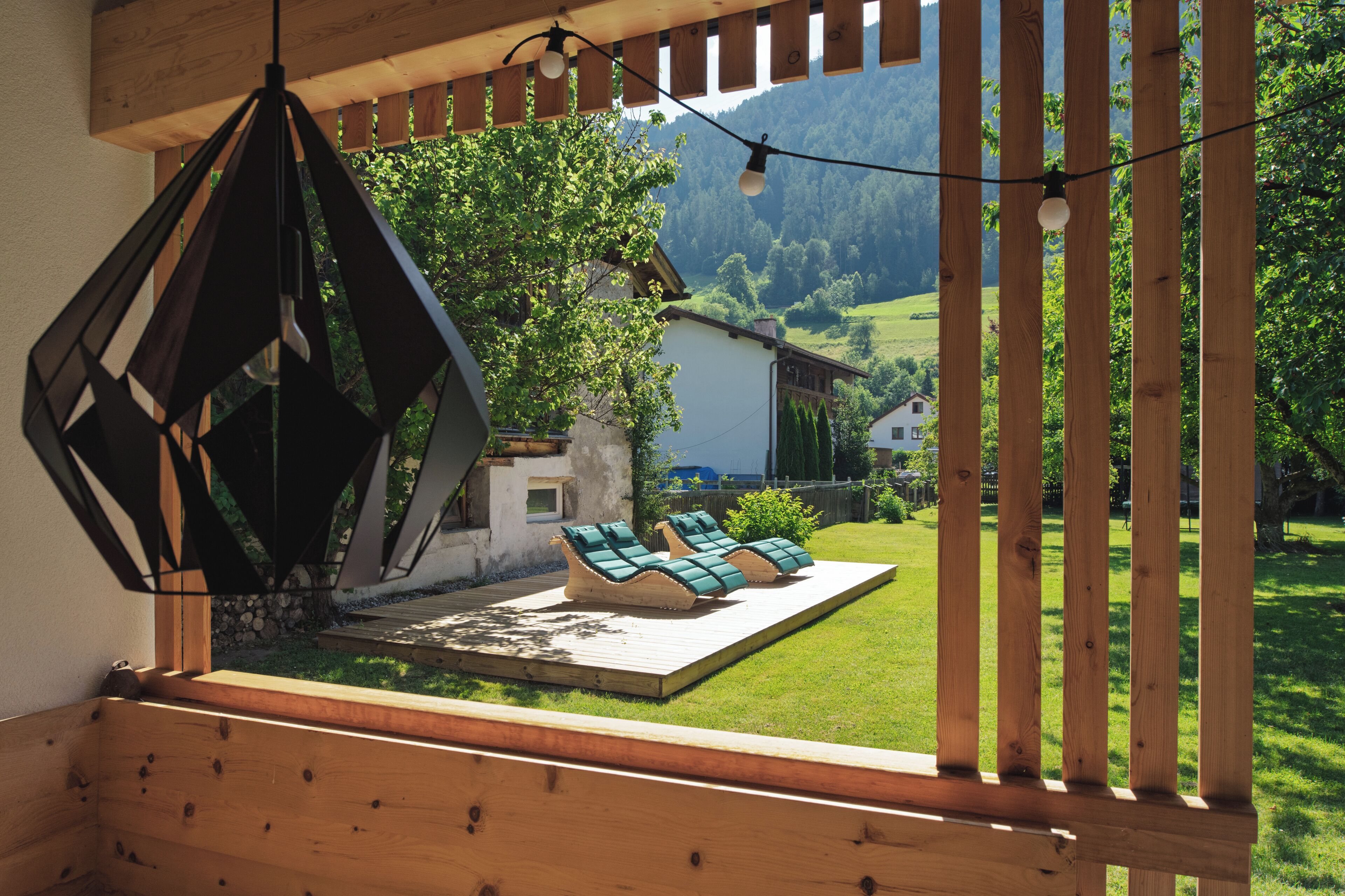Een houten terras met een geometrisch metalen zonnescherm, een houten pergola en uitzicht op een groen landschap met een huis.