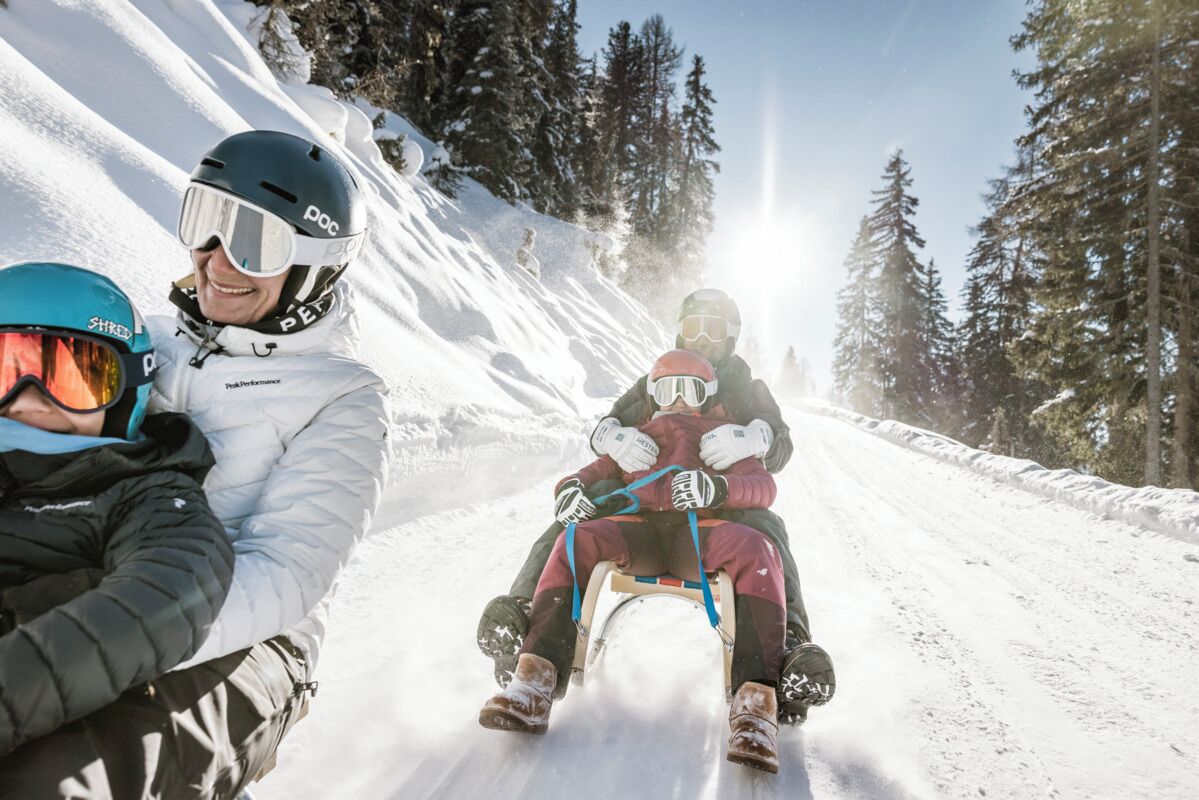 Une route de montagne enneigée, sur laquelle une personne tire une luge à l’aide d’une autre personne.