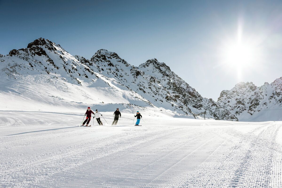 Ein verschneiter Skipisten mit Skifahrern, blauem Himmel und Bergen im Hintergrund.