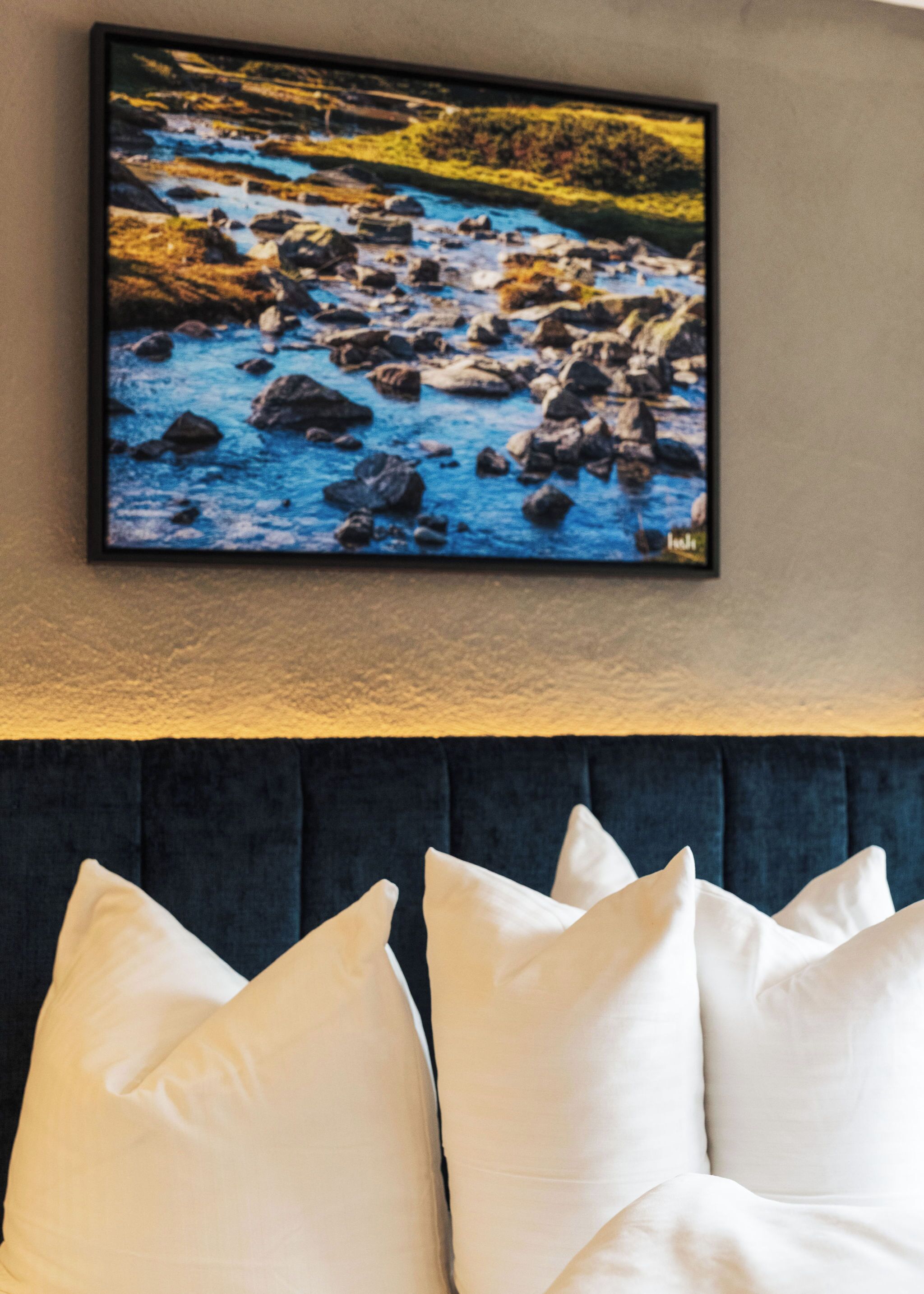 Une peinture représentant un paysage, encadrée, et qui montre une rivière avec des rochers. Elle est accrochée au mur au-dessus d’un lit agrémenté d’oreillers blancs.