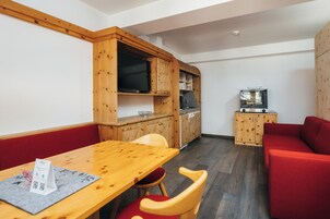 A compact room with a wooden dining table, red cushioned seating, a built-in TV, and a kitchenette with a stove.