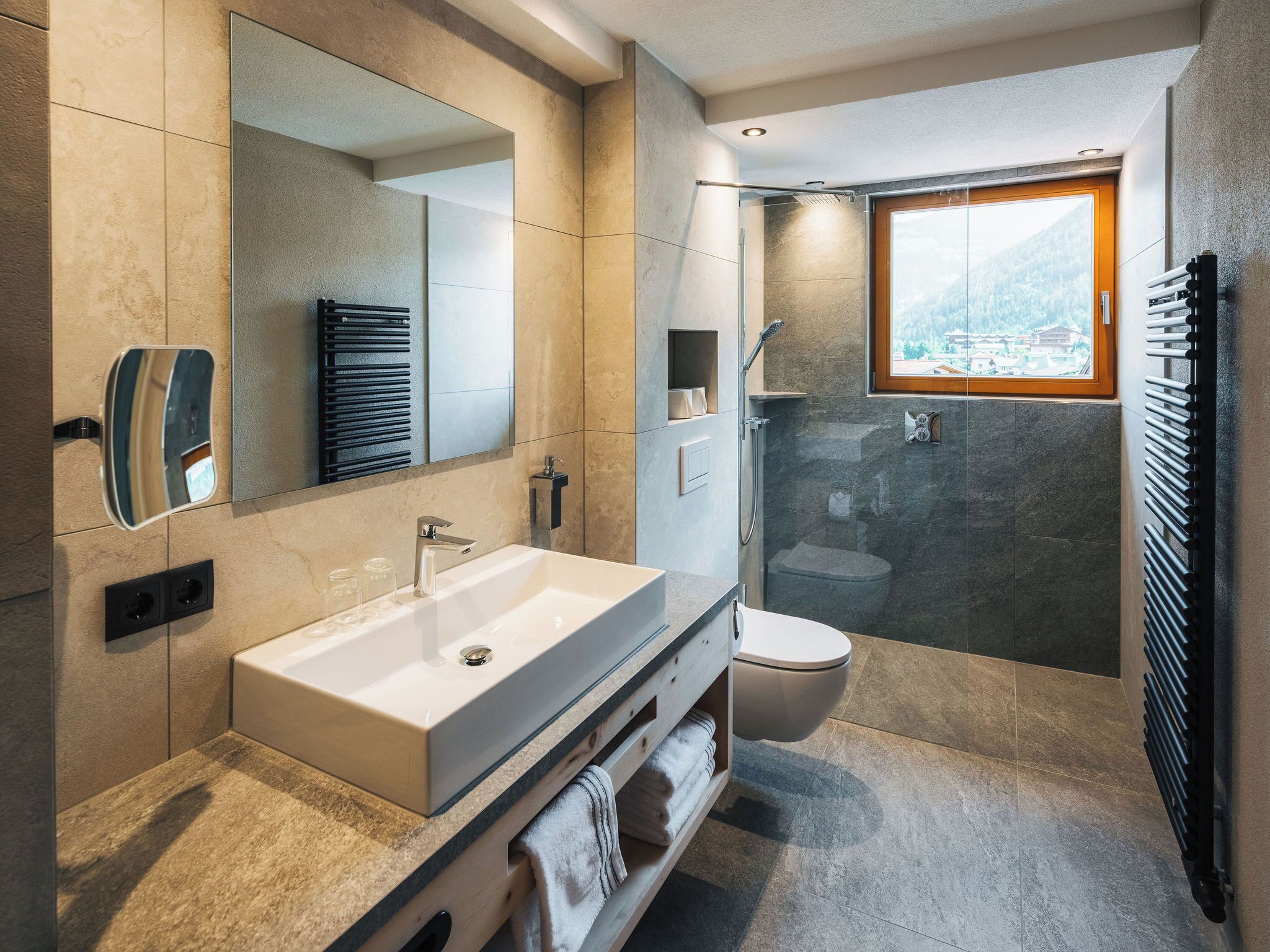 Ein modernes Badezimmer mit großem Spiegel, weißem Waschbecken, einer Dusche mit Glaswand und Blick ins Freie durch ein Fenster.