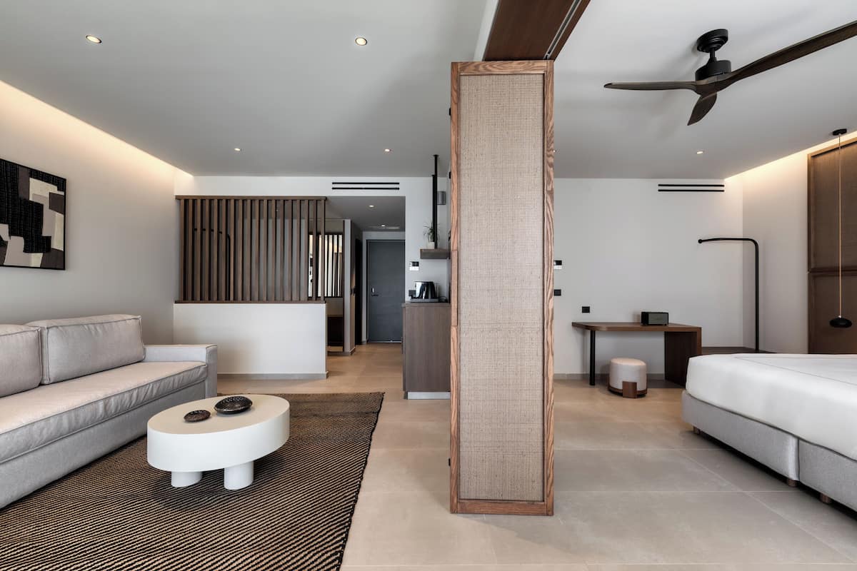 A modern hotel room with a sofa, a round coffee table, a bed, a desk, and a large window.