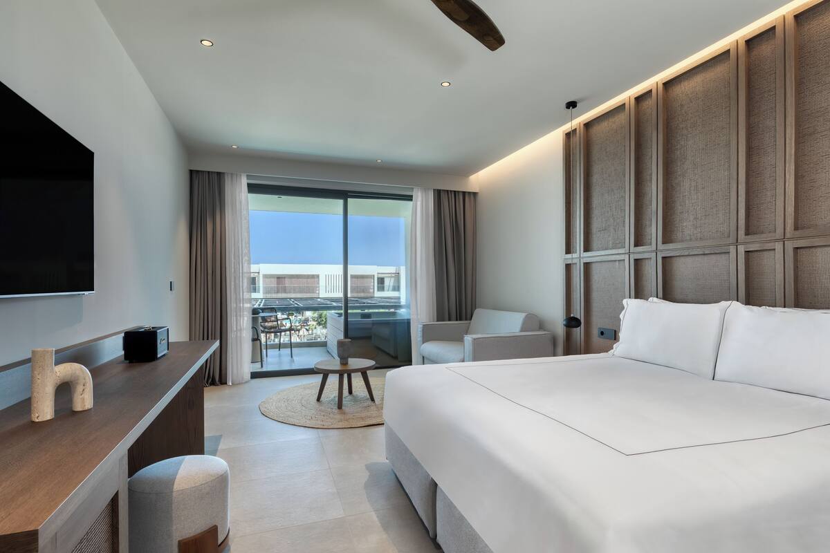 A modern hotel room with a large bed, a desk with a flat-screen TV, a seating area with a table and chairs, and a view of the city through large windows.